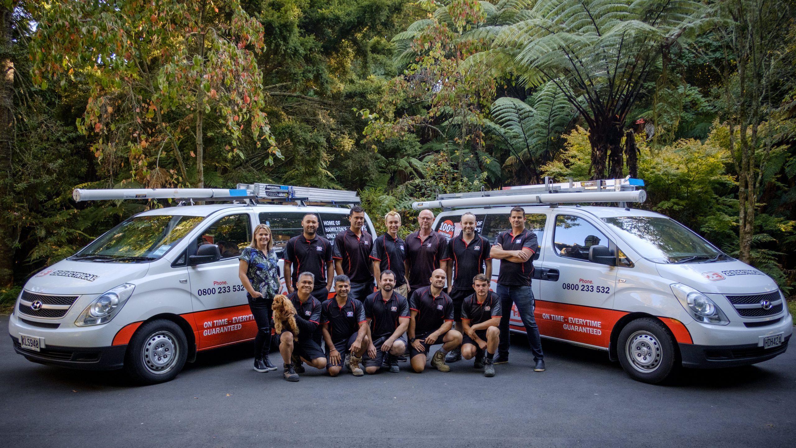 2E Electrical Hamilton team posing in front of branded vans in a lush outdoor setting