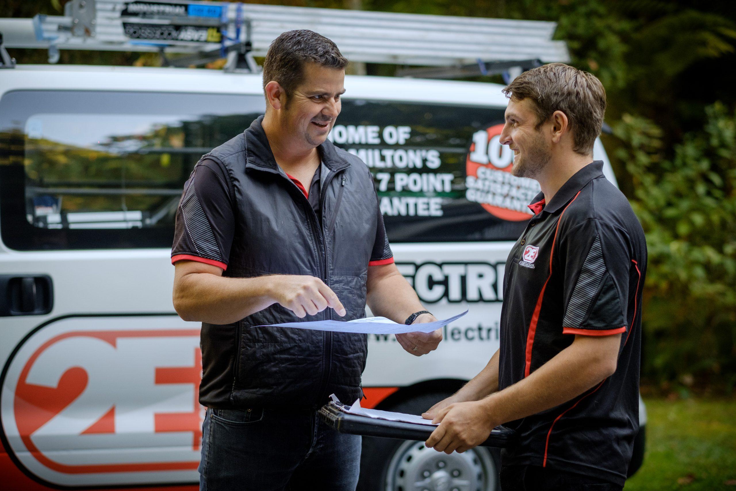 Two 2E Electrical team members discussing paperwork in front of a company van